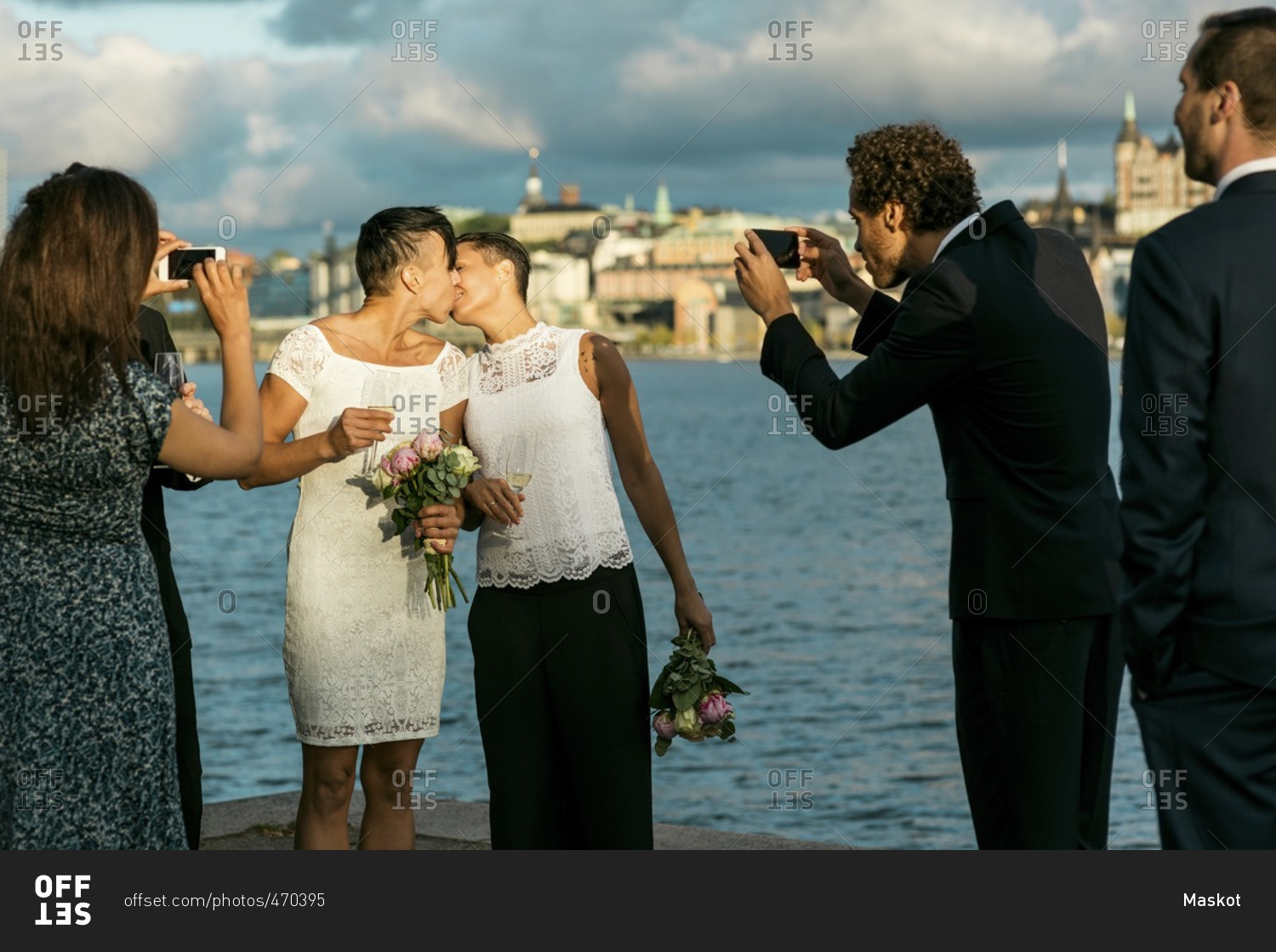 Happy man standing with friends while photographing lesbian couple kissing at wedding