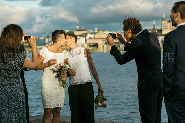 Happy man standing with friends while photographing lesbian couple kissing at wedding