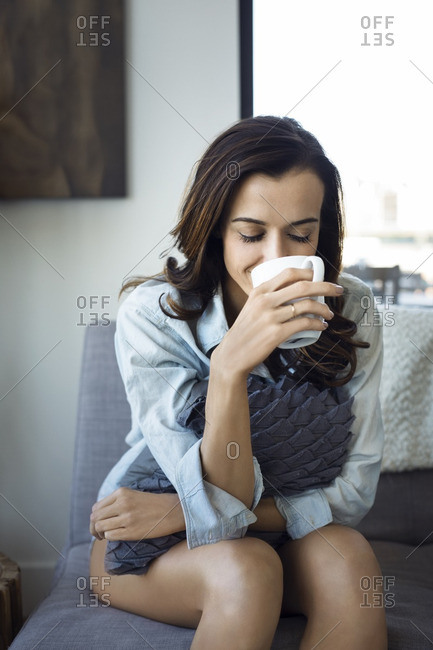 Woman holding cushion drinking while sitting on sofa