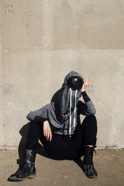 Woman in hooded shirt sitting on footpath against wall