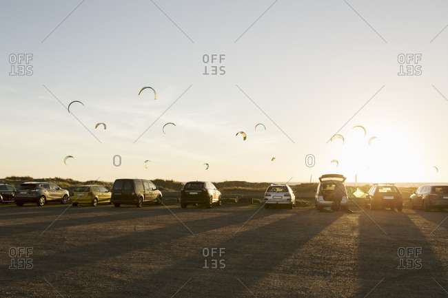 Cars parked on beach against parachutes in sky during sunset