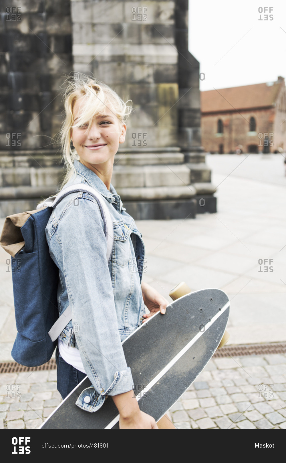 Smiling teenage girl holding skateboard while walking on cobbled street in city