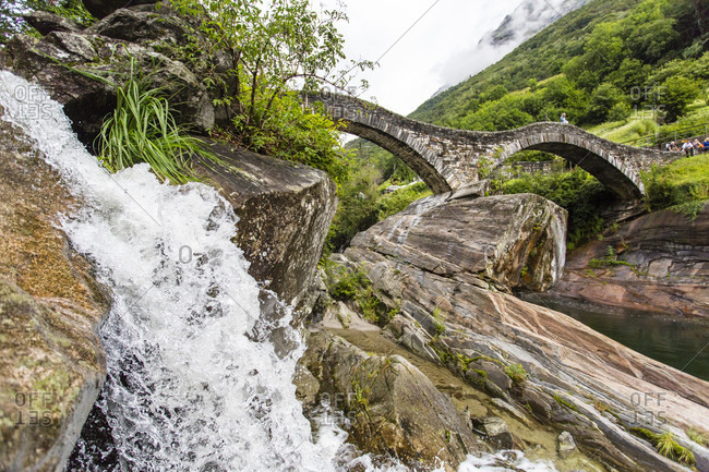Water Cascading Over Rocks With A Stone Bridge In The Background