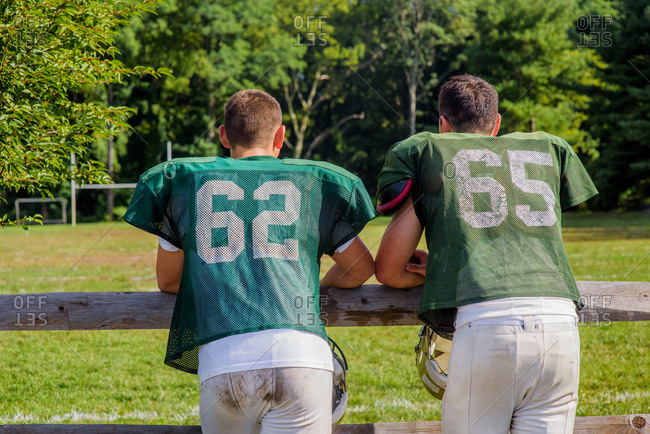 Rear view of two teenage male American football players leaning on playing field fence