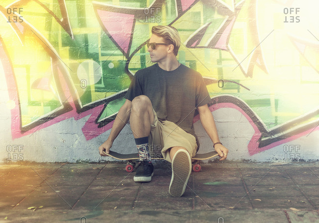 Young man sitting on skateboard, leaning against graffitied wall