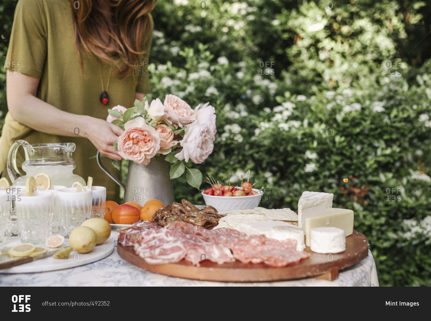 Woman standing at a table in a garden, a vase of pink roses, drinks, a bowl of cherries and a wooden board with cold cuts and cheese.