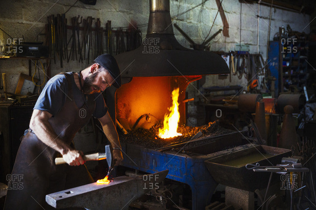 A blacksmith strikes a piece of red hot metal on an anvil with a hammer in a workshop.