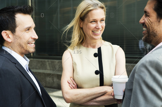 Businesswoman and two businessman standing outdoors, chatting and smiling.