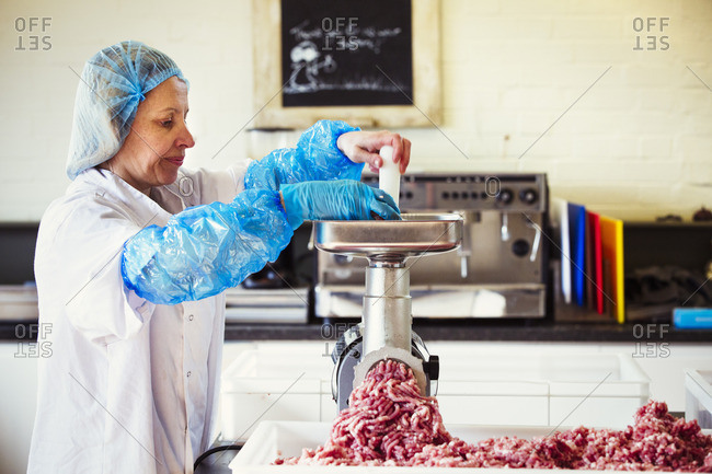 Woman working in a butchery, wearing protective clothes and gloves, putting minced meat into a meat grinder.