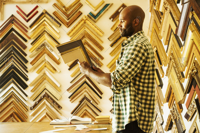 Man working at a picture framers, a large selection of frames on the walls.