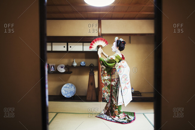 A woman dressed in the traditional geisha style, wearing a kimono and obi holding an open fan up, her kimono arranged on the floor.