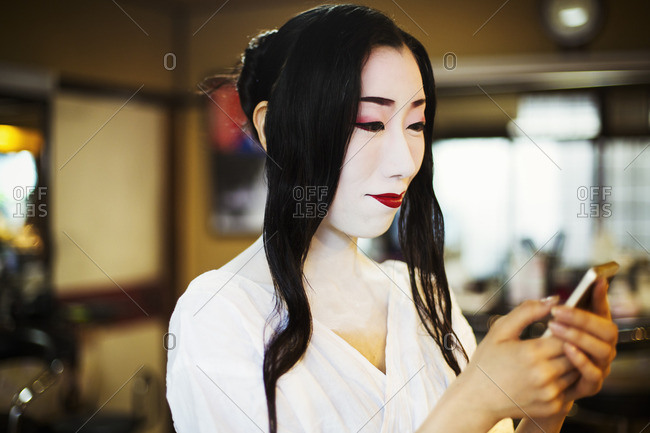 Geisha with long black hair and traditional white face make up  using a smart phone.