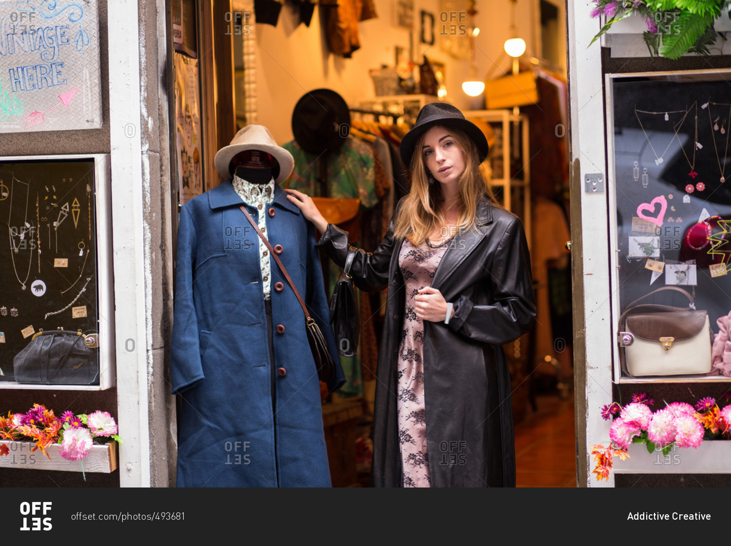 Young trendy girl posing near mannequin wearing coat in doorway of shop