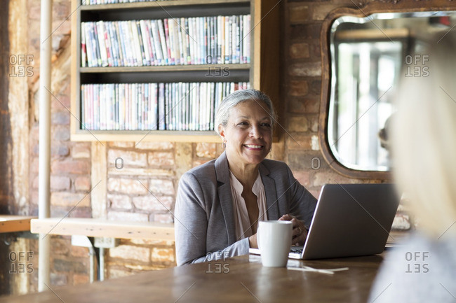 Smiling Woman Using Laptop While Sitting By Bookshelf At Cafe