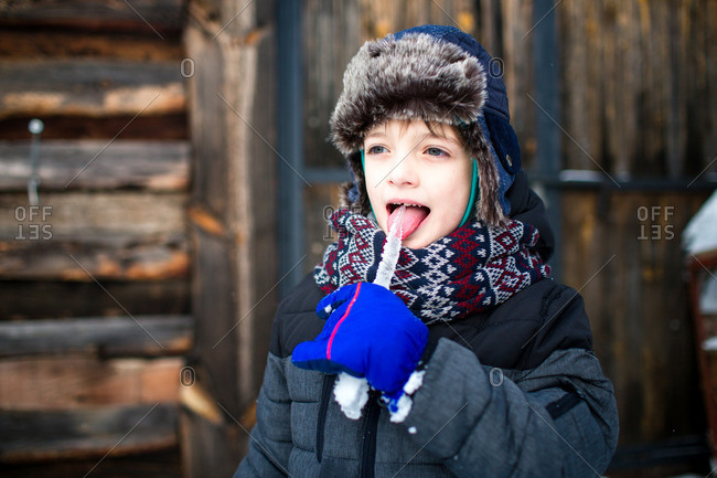 Happy child sticking out a tongue tasting an icicle with wooden cabin in the background