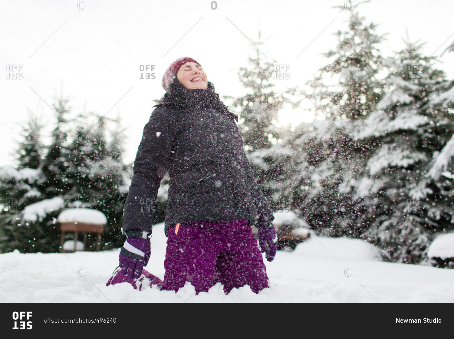 Laughing woman in warm winter clothes sitting on snowy ground having her face hit by snow