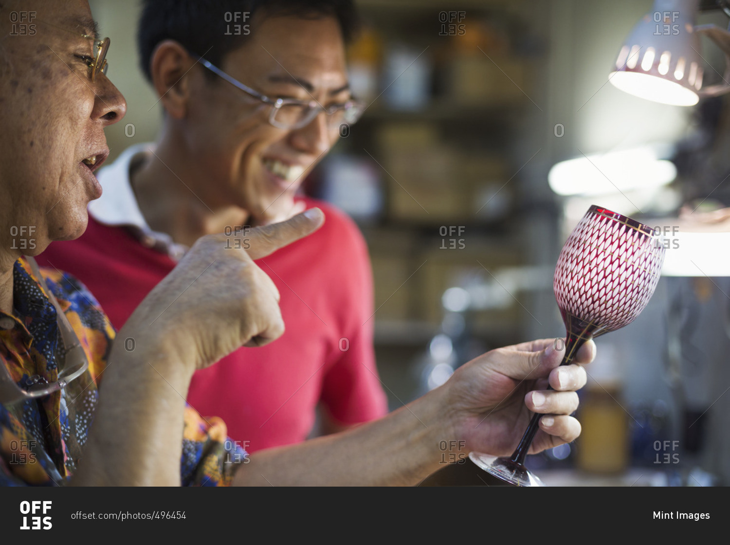 Father and son at work at a glass maker's studio workshop, inspecting a red cut glass wine glass.