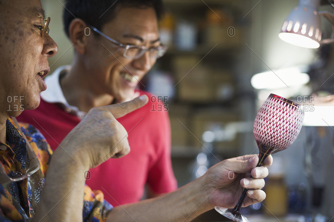 Father and son at work at a glass maker\'s studio workshop, inspecting a red cut glass wine glass.
