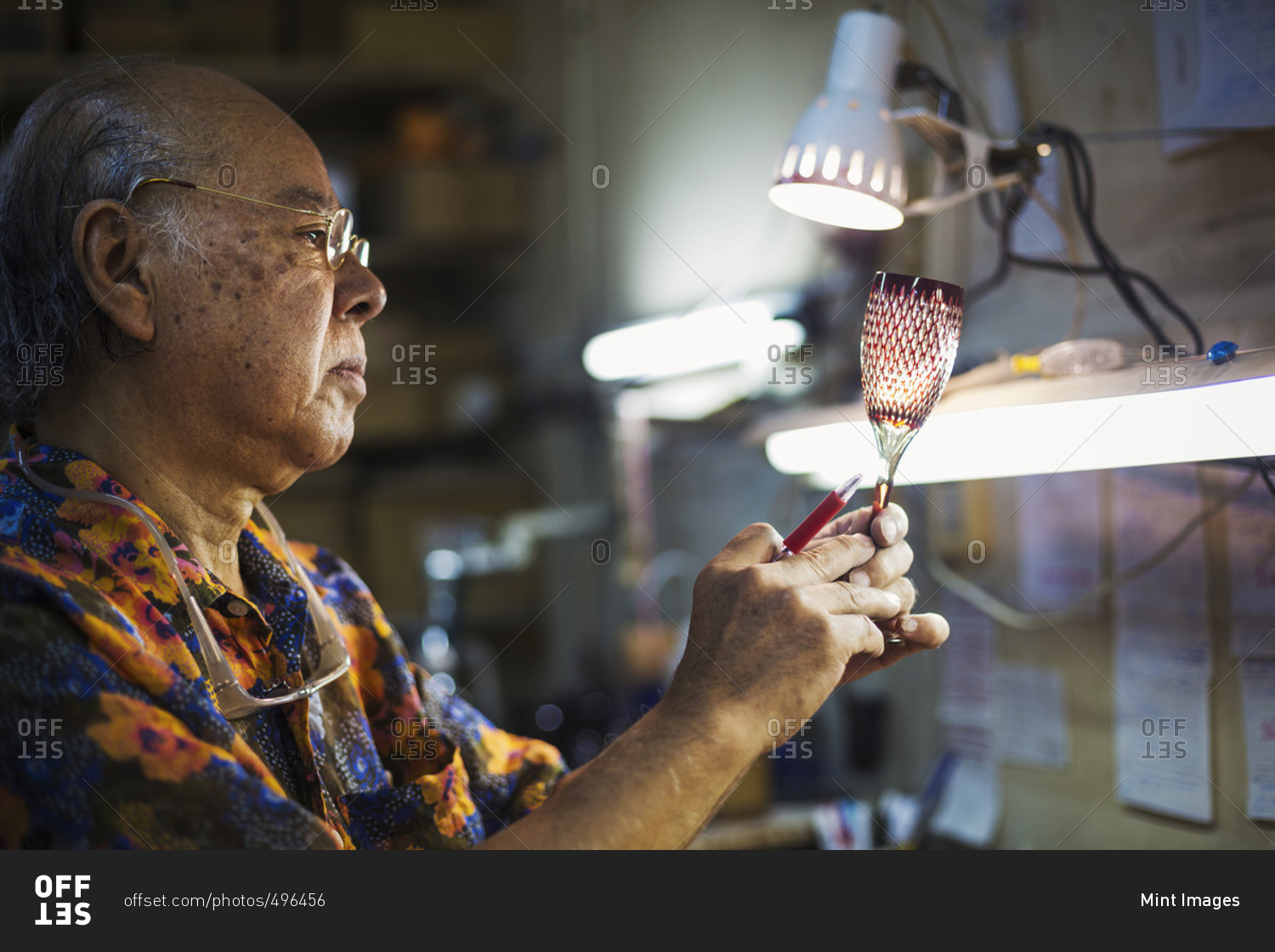 A senior craftsman at work in a glass maker\'s studio workshop, in inspecting red wine glass with cut glass decoration against the light.