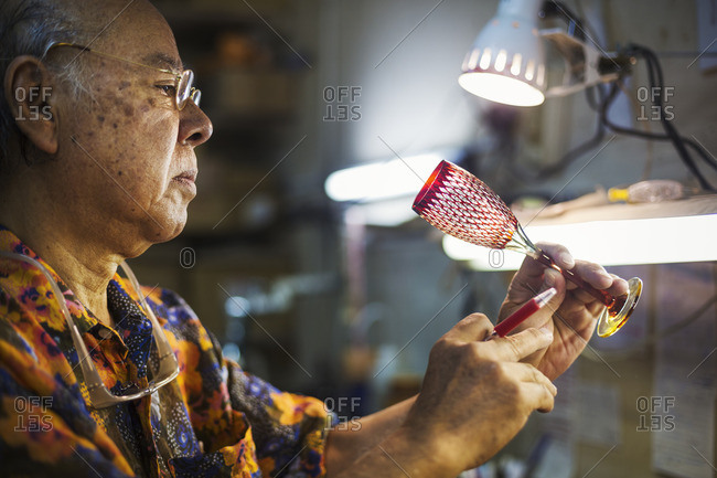 A senior craftsman at work in a glass maker's studio workshop, in inspecting red wine glass with cut glass decoration against the light.