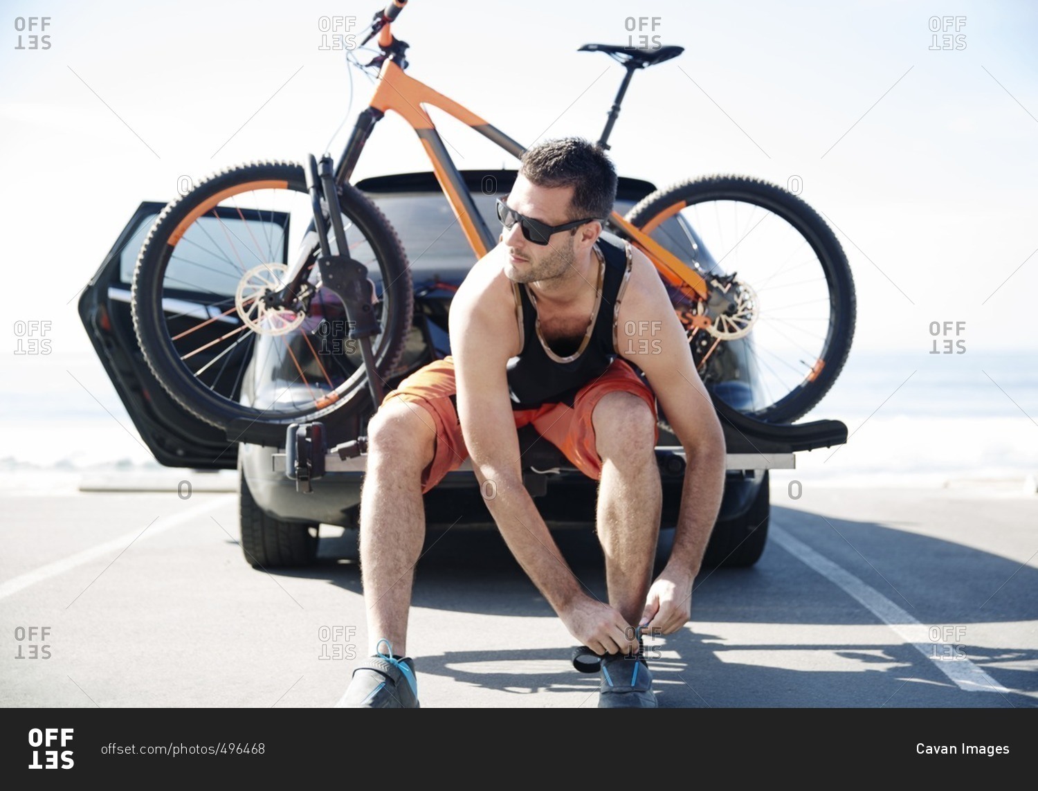 Man with bicycle tying shoelace while sitting on car against clear sky