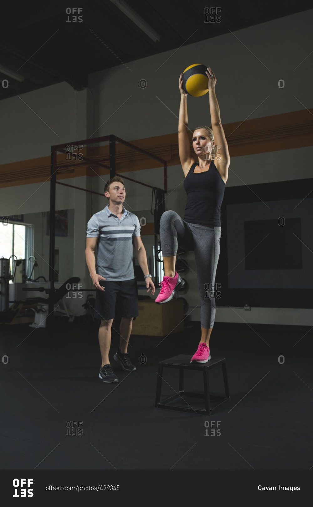 Trainer looking at woman holding fitness ball standing on stool in gym