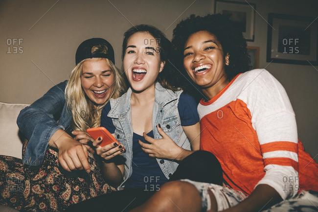 Cheerful woman holding smart phone while watching TV with friends during house party