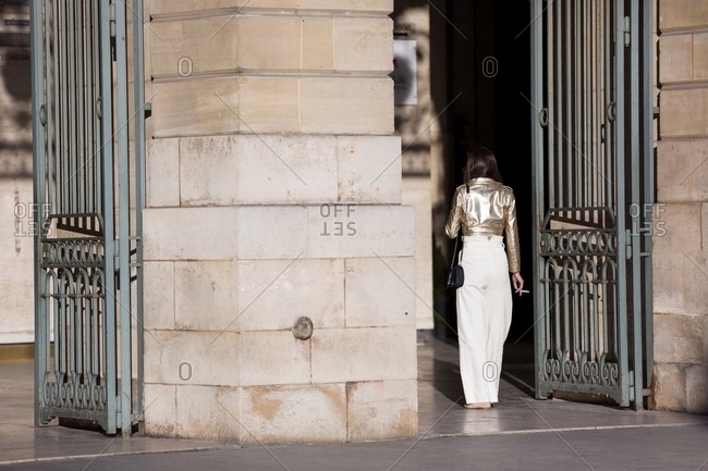 Back view of woman in white pants and gold leather jacket smoking a cigarette outside building