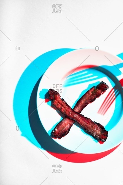 Two slices of bacon patterned in an X on a plate with two fork shadows.  The image is shot in a progressive modern style with two lights resulting in two shadows, a red and a blue.