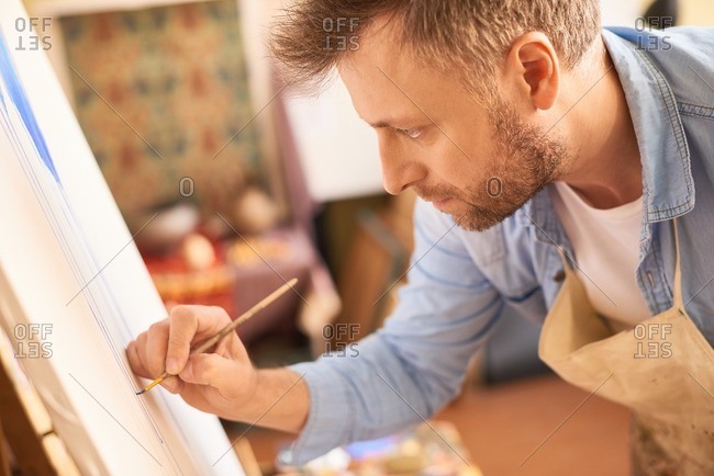 Inspired artist in studio. Concentrated bearded fair man in apron standing in front of easel and painting a picture with thin paintbrush