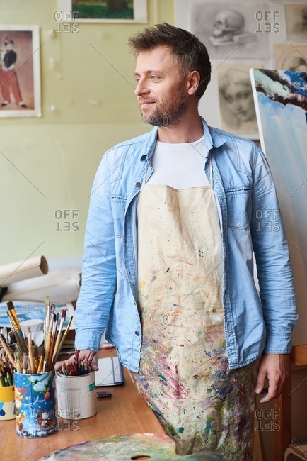 At art class. Dreamy middle-aged painter in stained apron standing in art studio, holding box of pencils and looking to the side