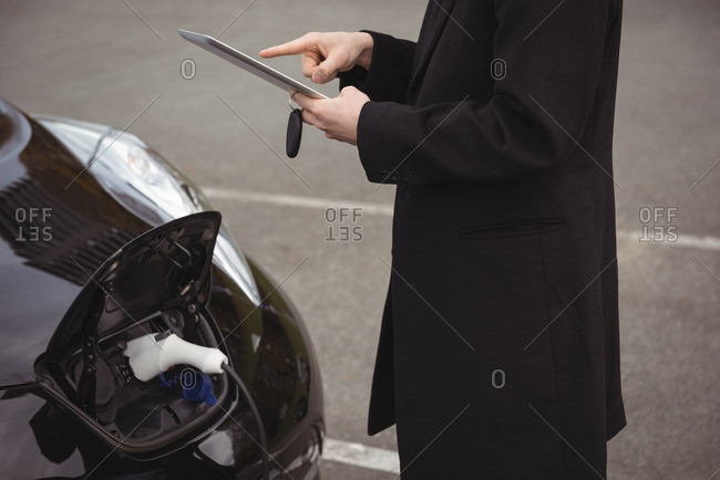 Man using digital tablet while charging electric car at electric vehicle charging station