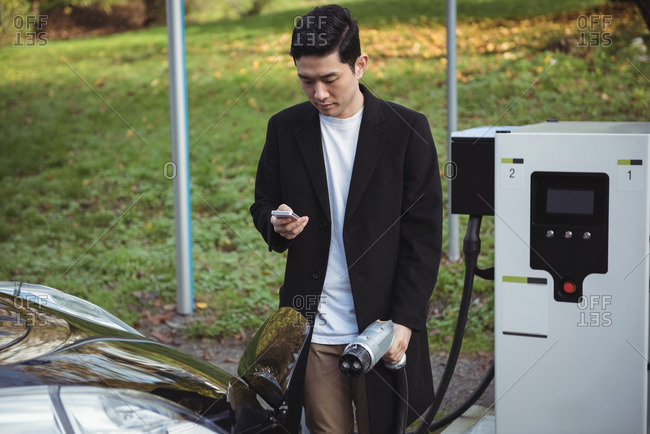 Man using mobile phone while charging car at electric vehicle charging station