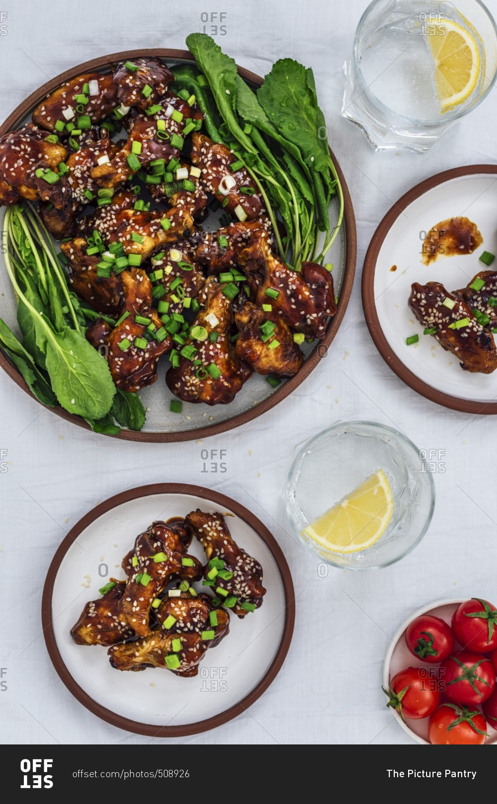 Oven-baked chicken wings with a sticky sauce and sesame seeds served with herbs on one large plate and two small plates accompanied by soda with lemon and cherry tomatoes.