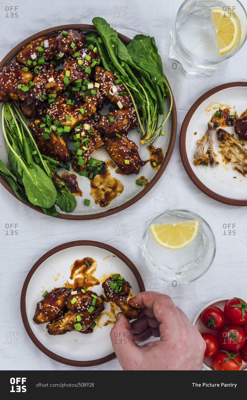 Oven-baked chicken wings with a sticky sauce and sesame seeds served with herbs on one large plate and two small plates accompanied by soda with lemon and cherry tomatoes. A man is grabbing one chicken wing.
