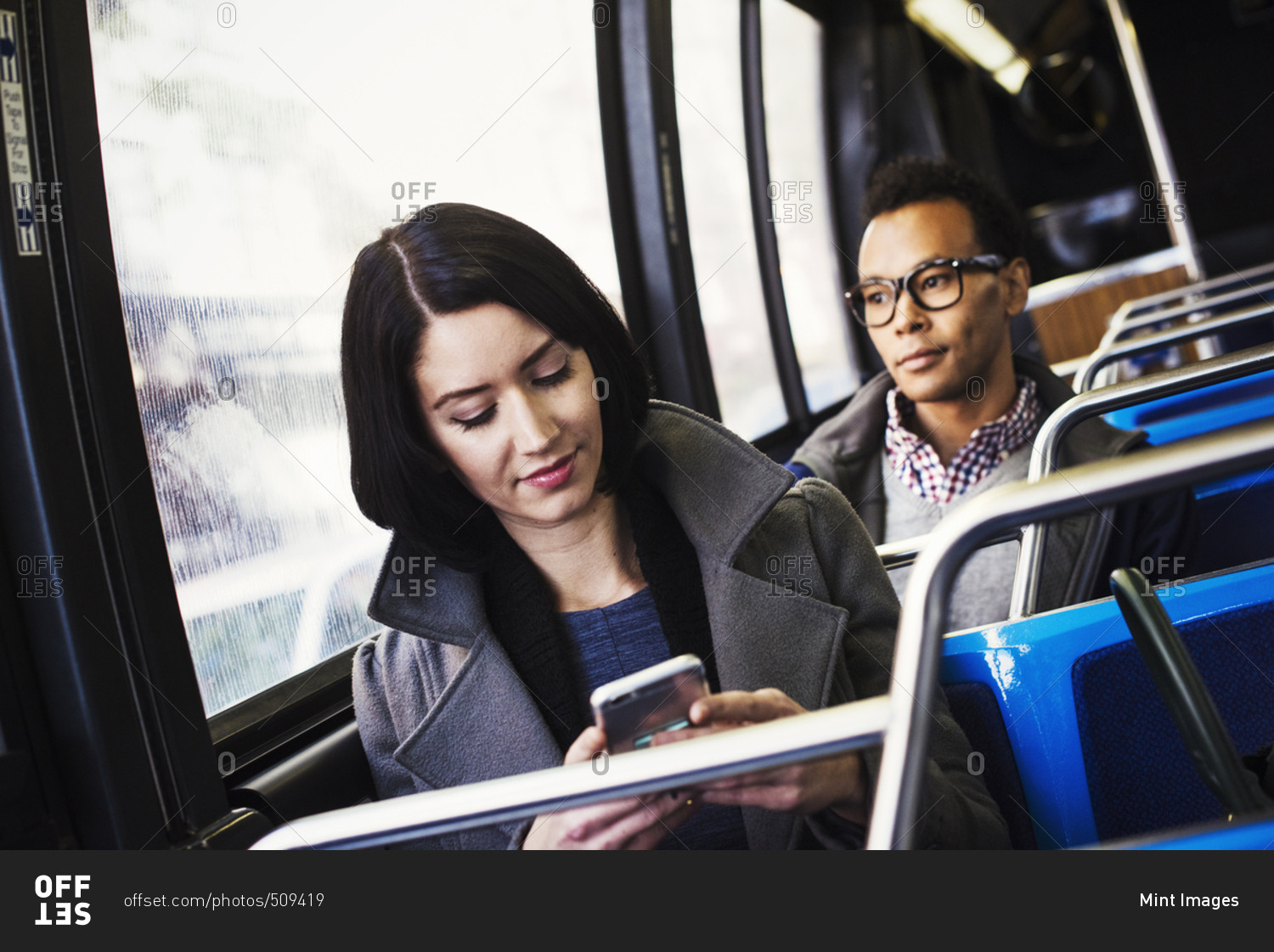 A young woman and a young man sitting on public transport, one looking at a cellphone and one looking away