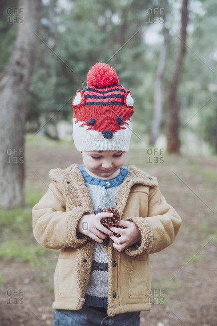Boy wearing wooly hat holding pine cone in forest