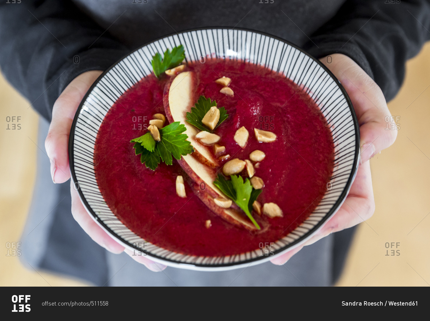 Girl\'s hands holding bowl of beetroot soup garnished with apple slices- peanuts and flat leaf parsley