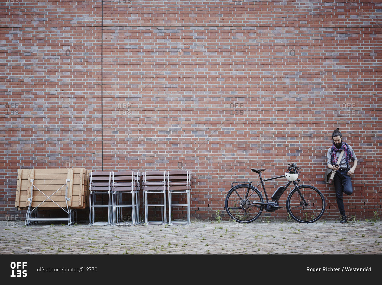 Man with electric bicycle leaning against brick wall looking at cell phone