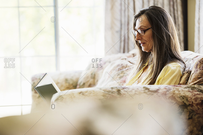 Senior woman with long brown hair sitting on a sofa, reading a book, wearing reading glasses.