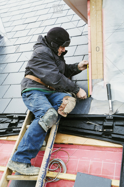 A man at the top of a ladder on a house roof, fixing tiles on a dormer roof.