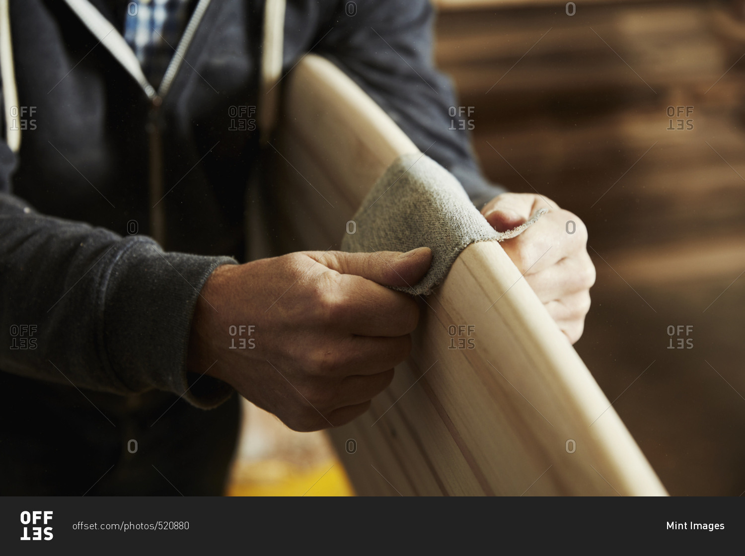 Man standing in a workshop sanding the rounded edge of a wooden surfboard.