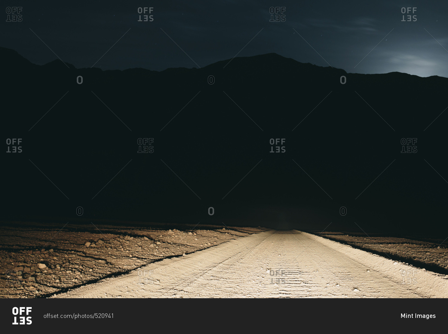Dirt road in desert illuminated by car headlights, Death Valley National Park, USA, with moonlight in distance.
