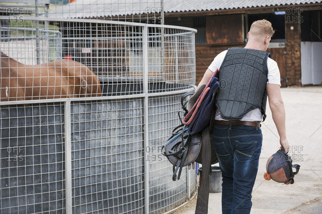 Rear view of a young man wearing a black body protector, and carrying riding gear at a riding stable.
