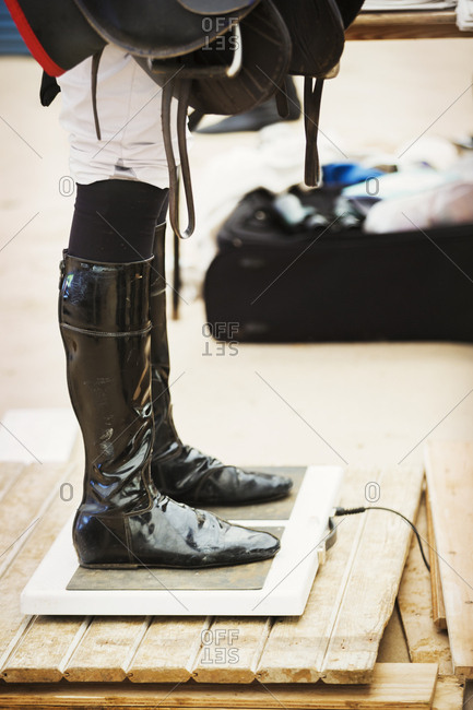 Rider wearing shiny black riding boots at the weigh in  on weighing scale, holding a saddle, before or after a race.