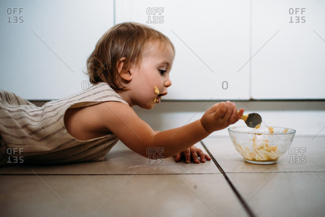 Toddler Sitting On Kitchen Floor Eating Hummus Out Of A Glass Bowl