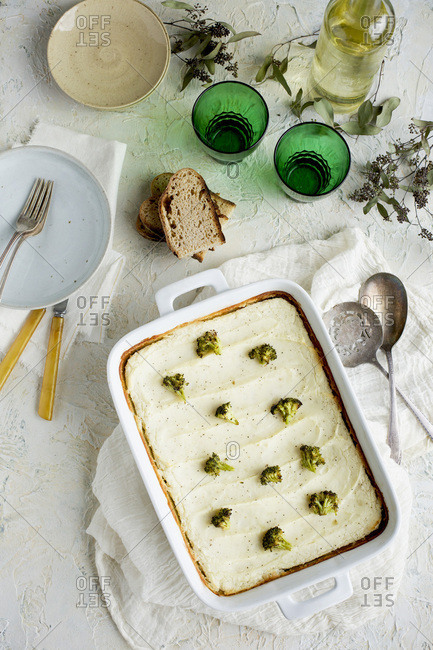 Black Lentil Charred Broccoli Shepherd's Pie served with bread and white wine. Photographed on white plaster background.