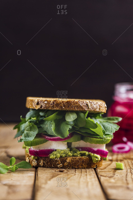 A springtime feta sandwich with avocado, pickled red onions and herbs like arugula, purslane and fresh mint served on a wooden board photographed from front view and a jar of pickled red onions accompanies on the background.