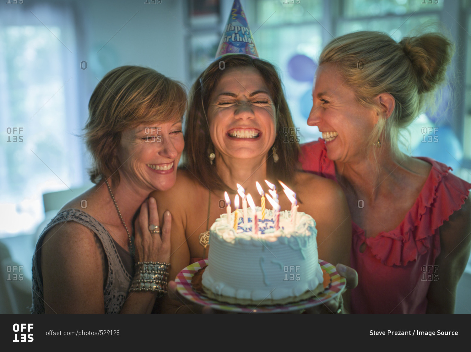 Mature woman holding birthday cake, making wish while two friends look on