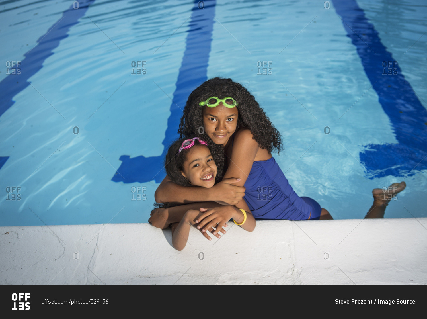 Girl in swimming pool with arms around little sister, looking at camera smiling